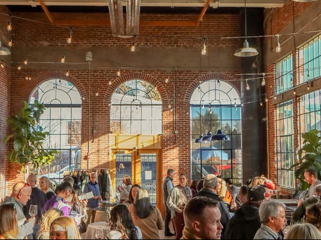 A lively indoor gathering in a brick-walled venue with tall arched windows, string lights, and guests mingling with drinks.