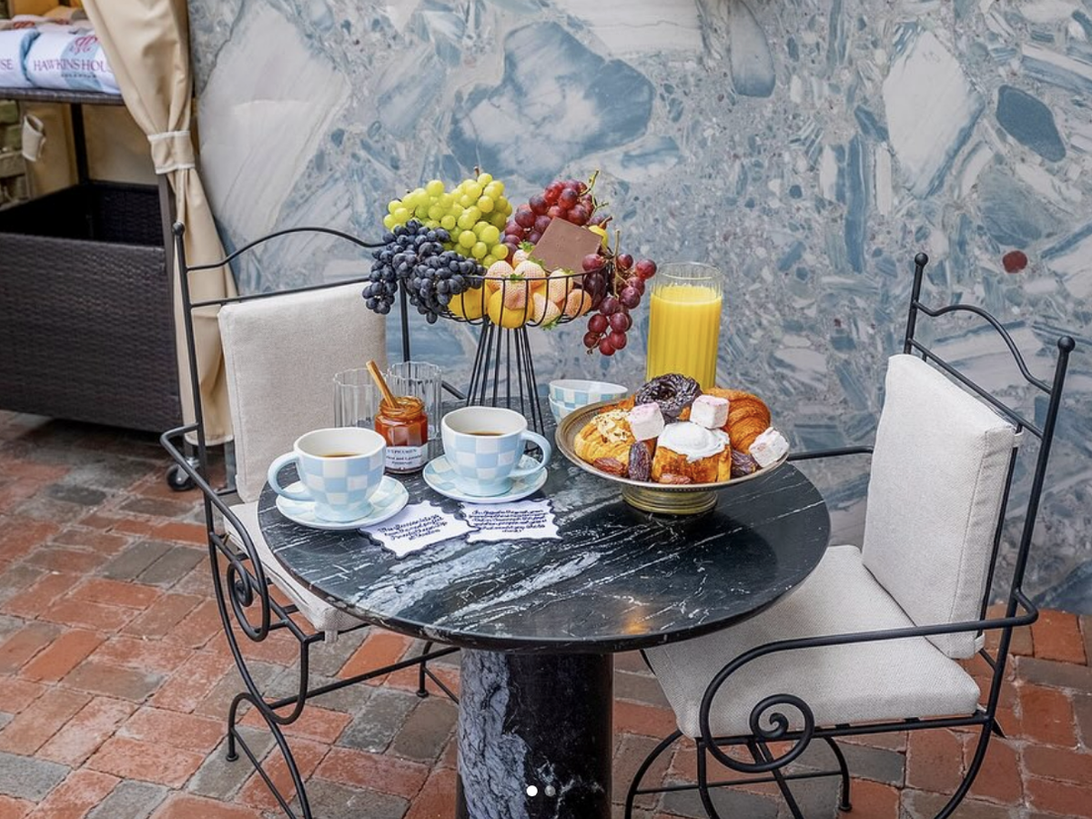 A stylish outdoor café setting with a black marble table holding two teacups, a glass of orange juice, a jar of honey, and a plate of assorted pastries. A tiered fruit basket with grapes and oranges adds color, set against a blue marbled wall on a brick patio