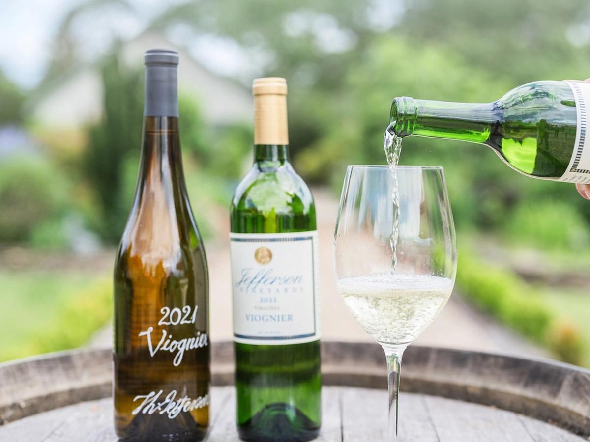 A crisp glass of white wine is poured beside bottles of Viognier at Jefferson Vineyards. The outdoor garden setting and bright natural light celebrate the elegance and freshness of Monticello’s wines.
