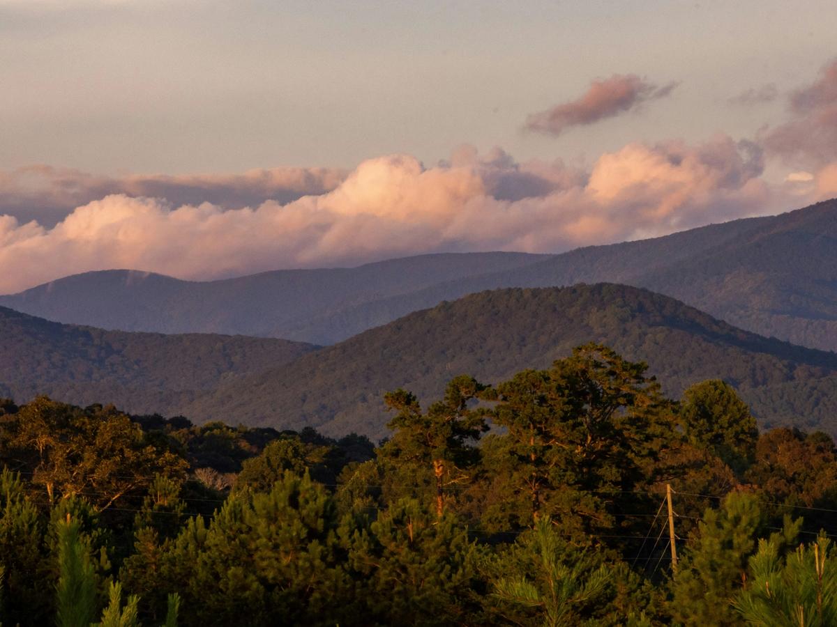 Layers of forested mountains stretch into the distance beneath a sky filled with soft, pink-tinged clouds at sunset.