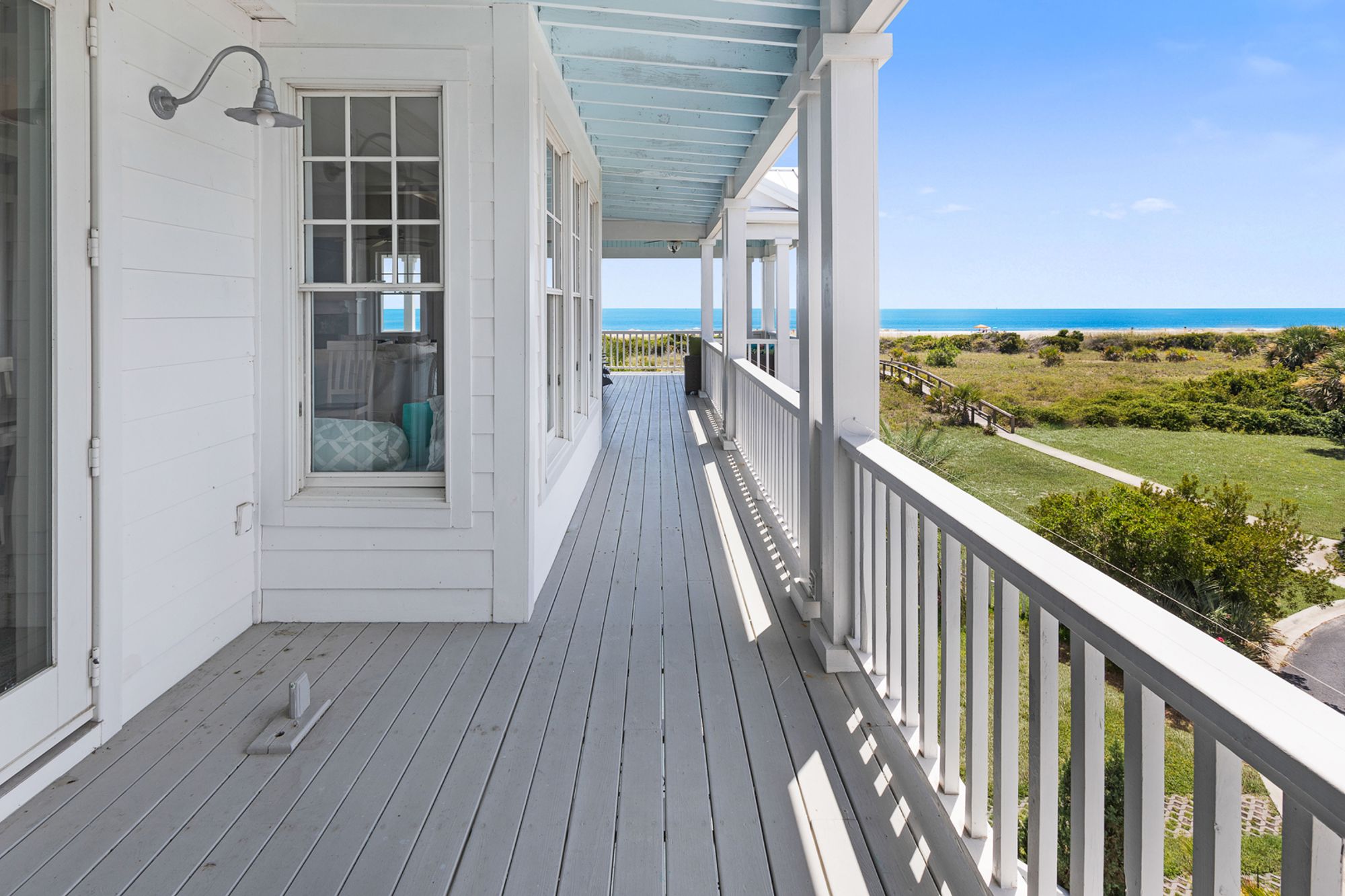 Wraparound beach house porch with white railings and light blue ceiling, offering views of a lush dune path leading to the ocean under a clear blue sky.        Ask ChatGPT