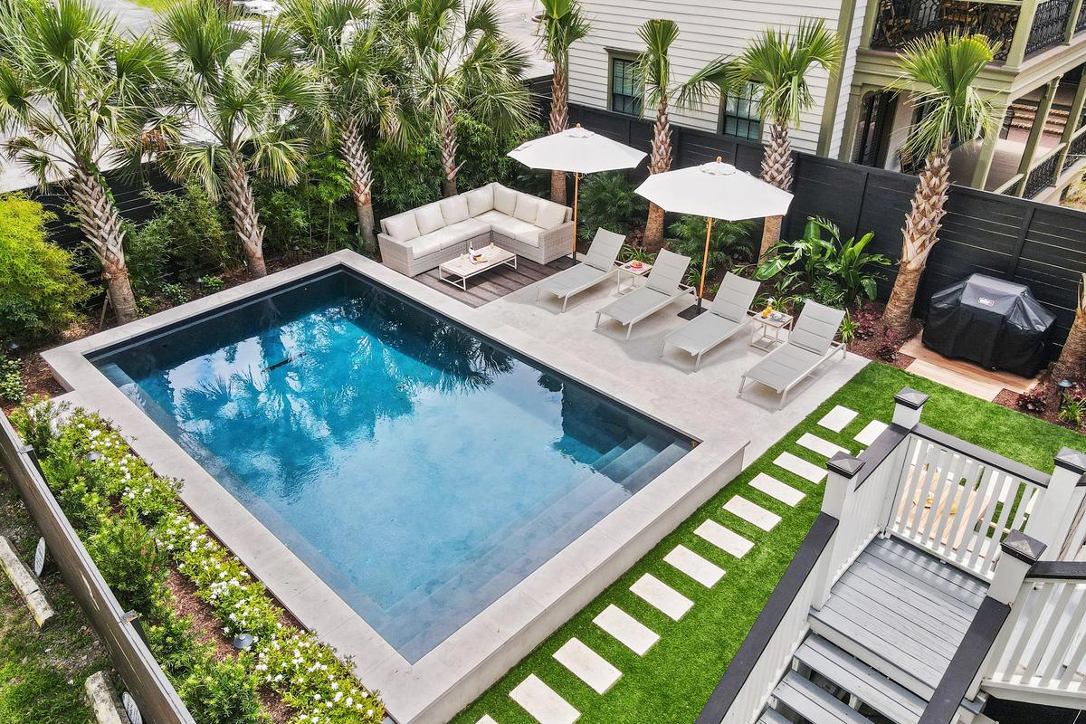 A modern backyard oasis features a rectangular pool surrounded by palm trees, cushioned lounge chairs with umbrellas, a corner outdoor sofa, and a barbecue grill area.