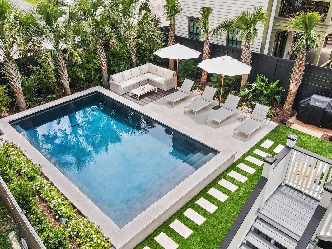 A modern backyard oasis features a rectangular pool surrounded by palm trees, cushioned lounge chairs with umbrellas, a corner outdoor sofa, and a barbecue grill area.