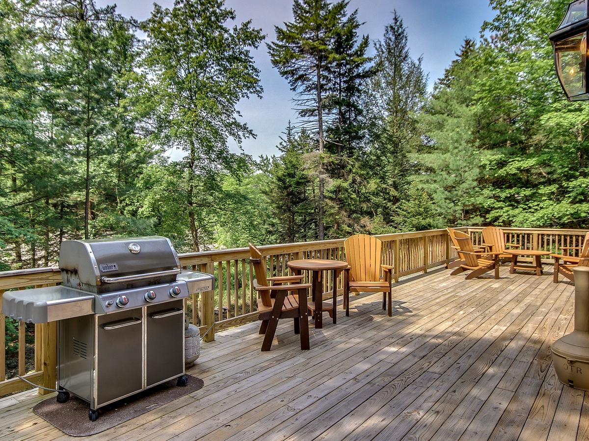 Spacious wooden deck with a stainless steel grill, Adirondack chairs, and forest views, perfect for outdoor dining and relaxation.