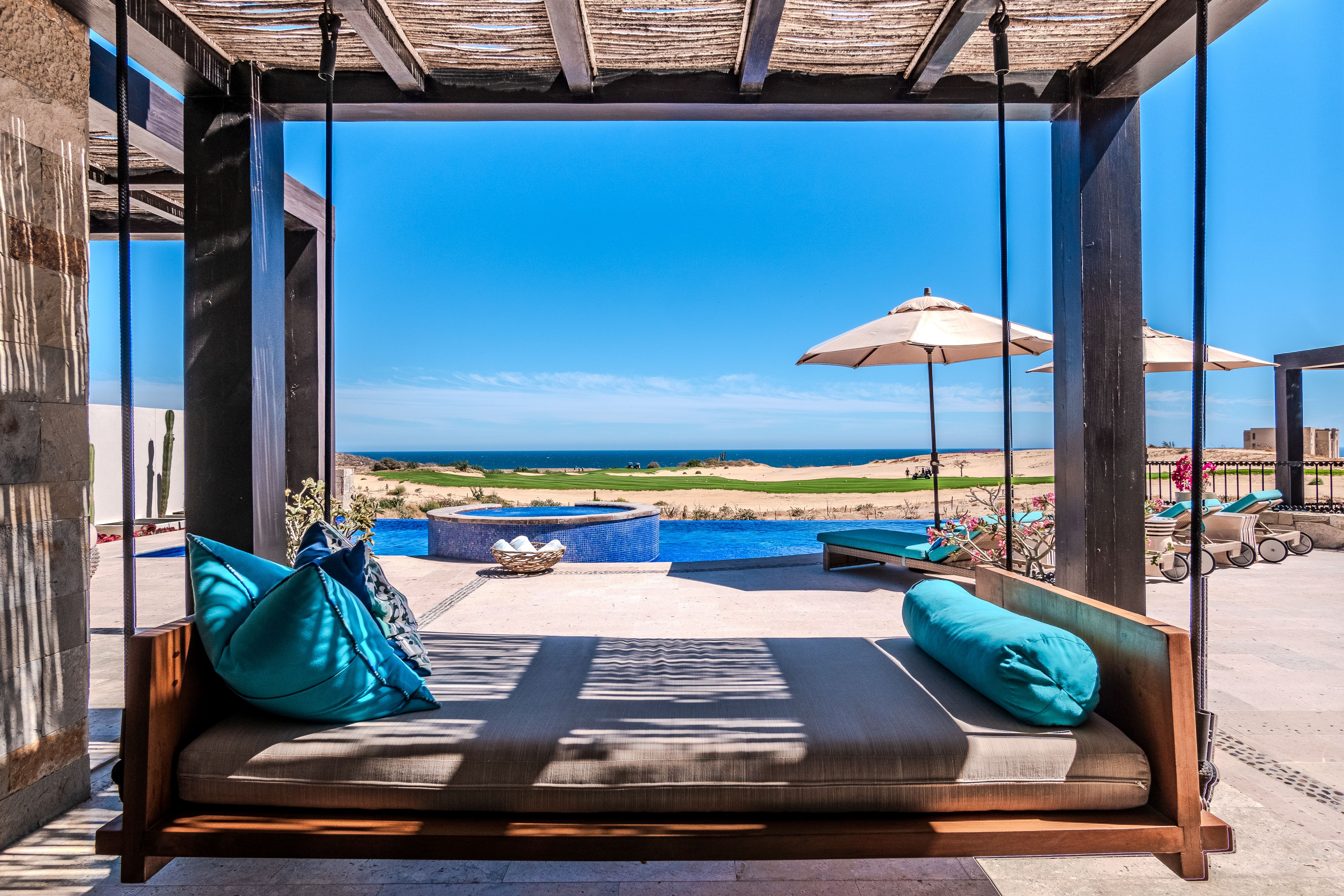 Luxurious shaded daybed swings beneath a wooden pergola, facing an infinity pool, hot tub, and sun loungers with umbrellas overlooking a desert coastline and the ocean.
