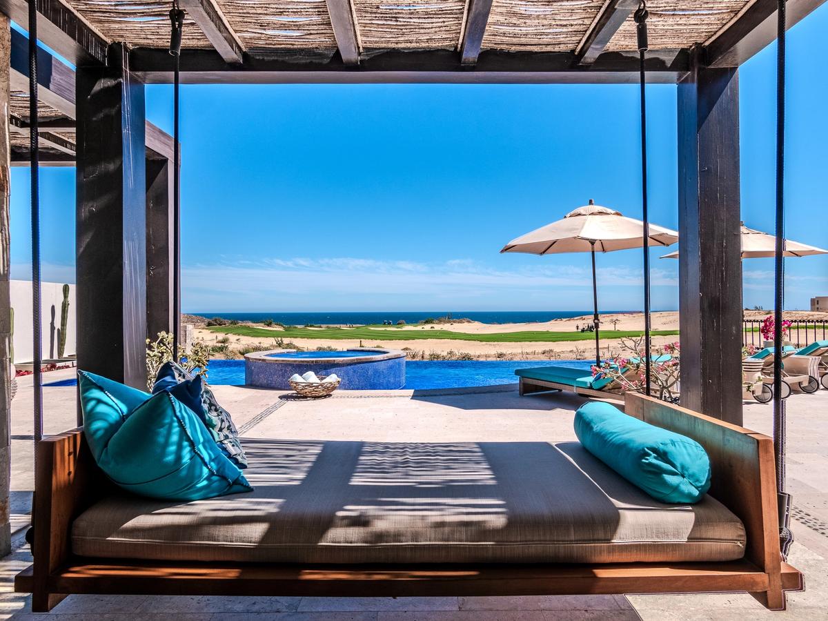Luxurious shaded daybed swings beneath a wooden pergola, facing an infinity pool, hot tub, and sun loungers with umbrellas overlooking a desert coastline and the ocean.