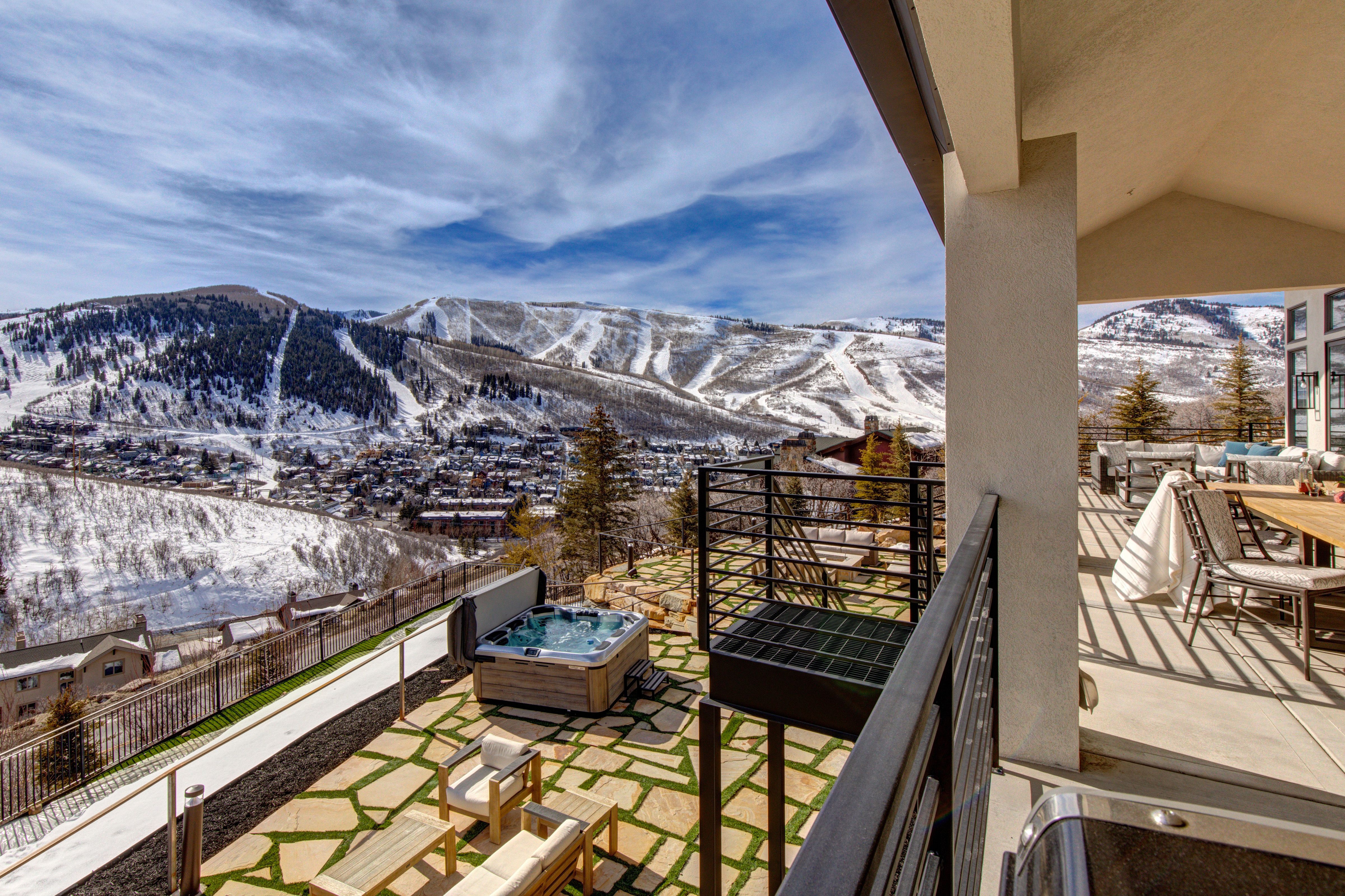 A luxury mountain home terrace featuring a hot tub, lounge seating, and dining area, with panoramic views of snow-covered ski slopes and a charming alpine town below.