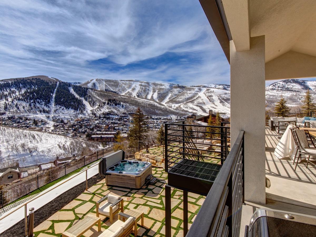 A luxury mountain home terrace featuring a hot tub, lounge seating, and dining area, with panoramic views of snow-covered ski slopes and a charming alpine town below.