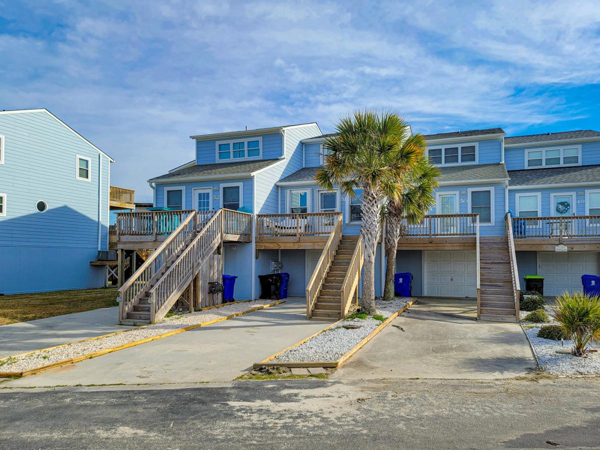 A row of coastal blue townhomes with wooden staircases leading up to elevated decks, surrounded by palm trees, white gravel landscaping, and driveways with recycling bins.