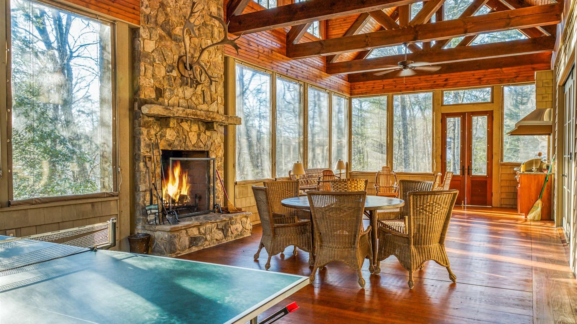 A spacious sunroom with a stone fireplace and large windows creates a warm gathering space filled with natural light. The dining table and game area make this cabin perfect for family meals and indoor fun.