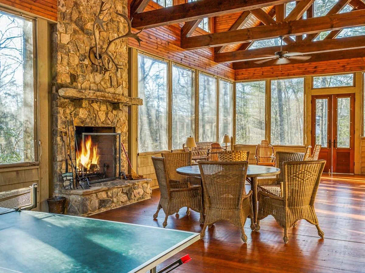 A spacious sunroom with a stone fireplace and large windows creates a warm gathering space filled with natural light. The dining table and game area make this cabin perfect for family meals and indoor fun.