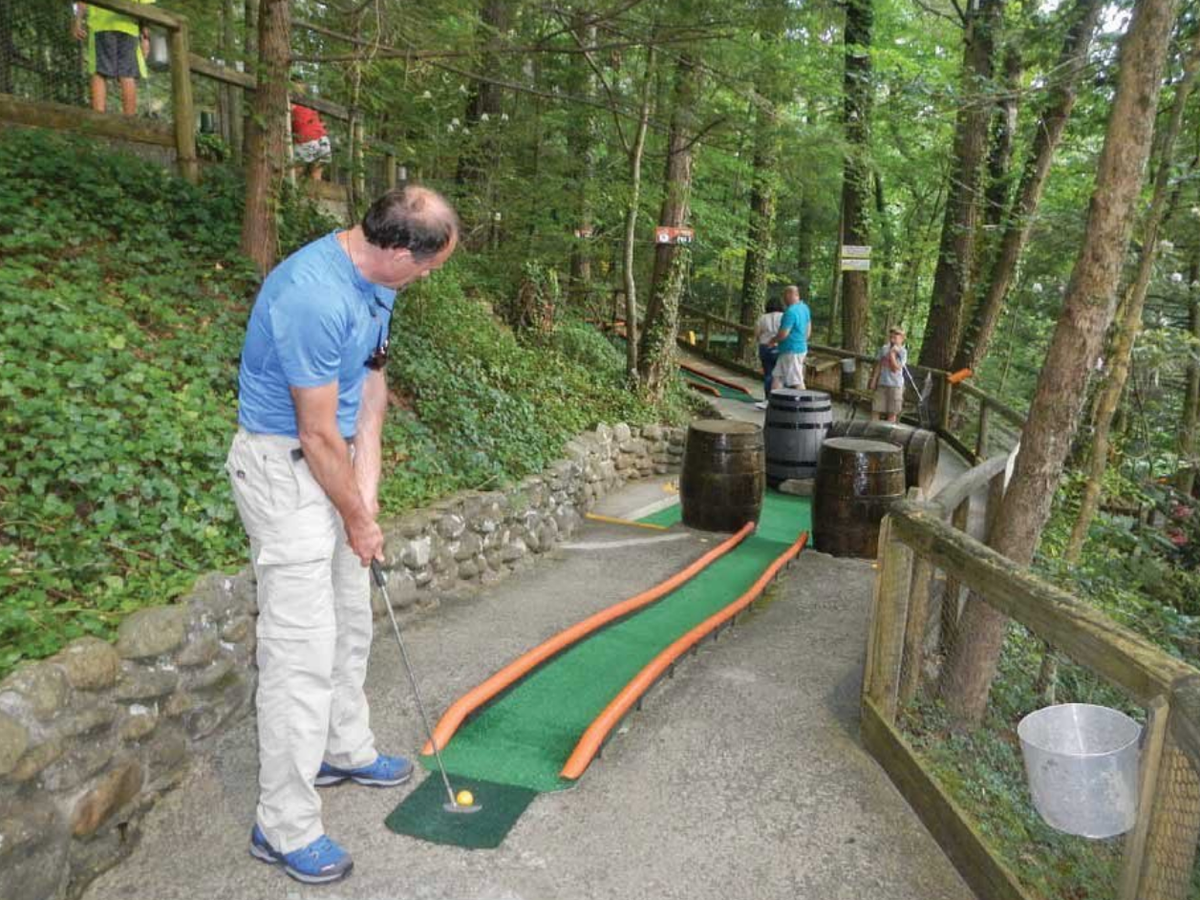 A man lines up a putt on a quirky outdoor mini-golf course at Hillbilly Golf, with wooden barrels serving as obstacles and lush forest surrounding the players.