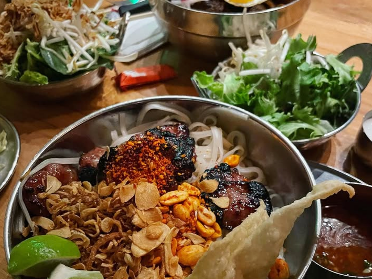 Colorful bowls of Vietnamese noodle dishes topped with grilled meat, peanuts, fried garlic, lime, and fresh herbs, served with sides of greens and dipping sauces.