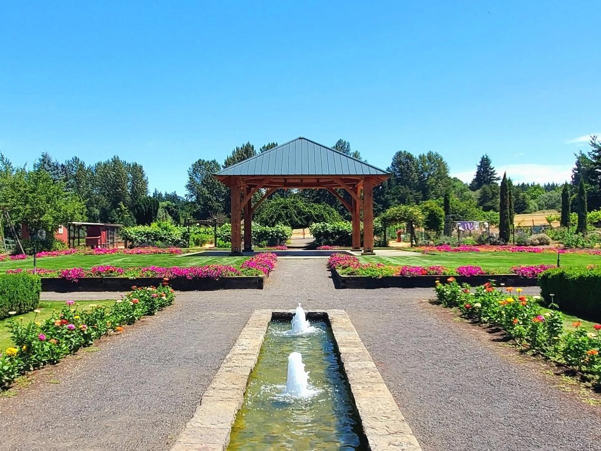 A long garden walkway leads to a wooden pavilion surrounded by colorful flower beds and neat hedges. Two small fountains add movement to the calm, sunny space. It’s a relaxing spot that shows the beauty of The Oregon Garden.