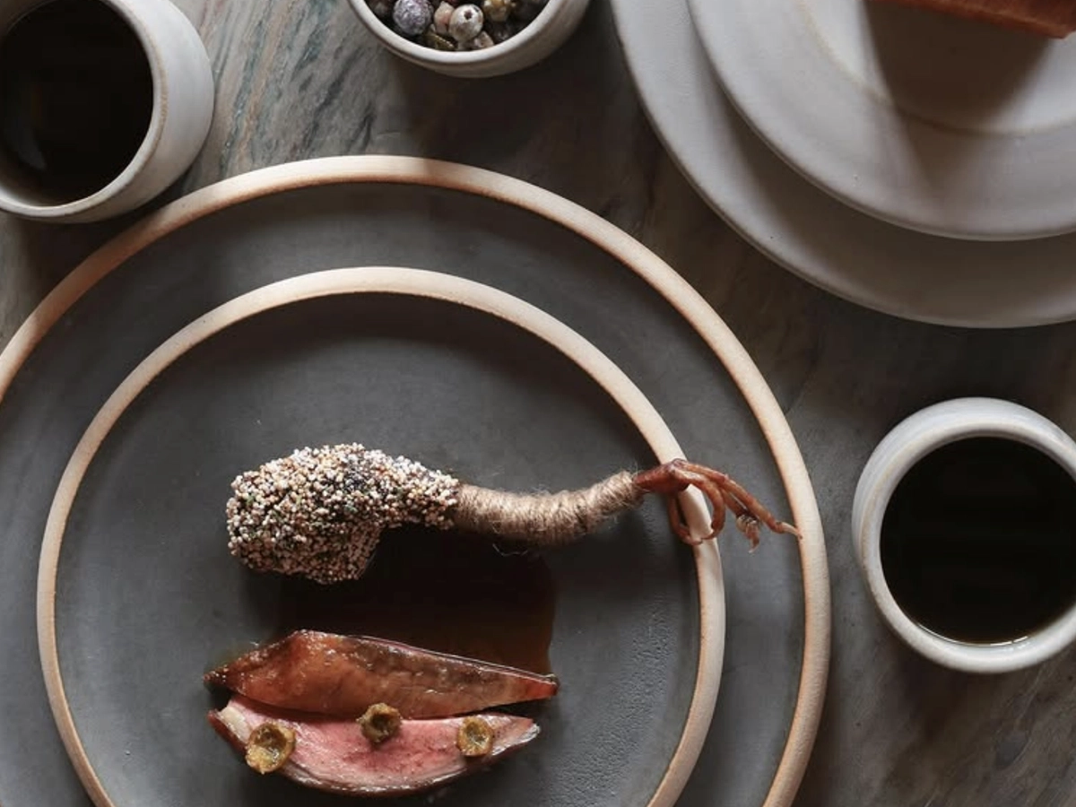 Artistic fine dining presentation featuring slices of seared meat with sauce and a garnished leg piece, served on minimalist ceramic plates with coffee and small accompaniments.