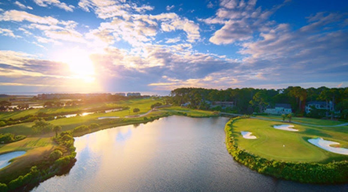 An aerial sunset view of the Robert Trent Jones Course at Palmetto Dunes on Hilton Head Island reveals a waterfront green complex surrounded by a large lagoon, with the Atlantic Ocean and resort skyline glowing in the background