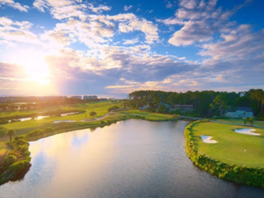 An aerial sunset view of the Robert Trent Jones Course at Palmetto Dunes on Hilton Head Island reveals a waterfront green complex surrounded by a large lagoon, with the Atlantic Ocean and resort skyline glowing in the background