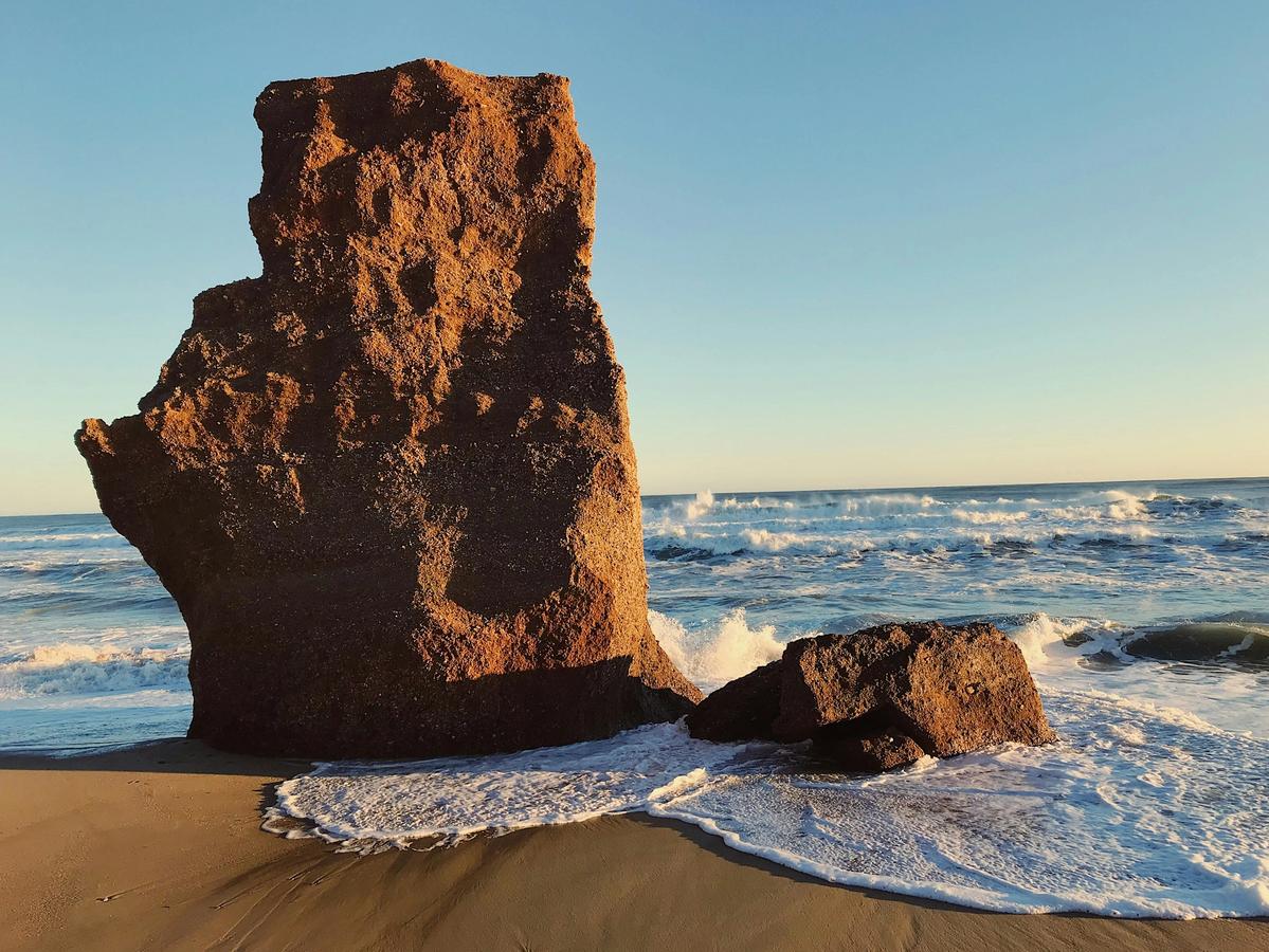 A dramatic sea stack towers over the shoreline as waves crash around its base, bathed in golden light from the setting sun.