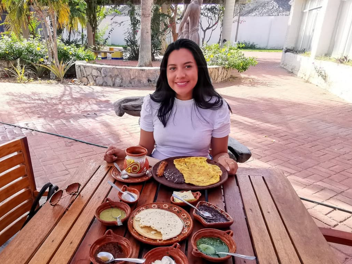 A woman sits at a wooden outdoor table smiling, enjoying a traditional Mexican breakfast with an omelet, black beans, plantain, handmade tortillas, salsas, and a mug of coffee in a sunlit courtyard setting.