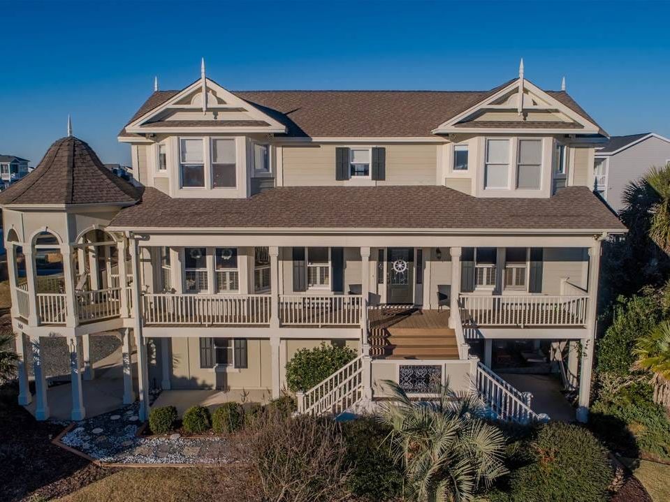 A large, coastal-style home with Victorian-inspired architecture, multiple gables, and a wraparound porch surrounded by palm trees and manicured landscaping.