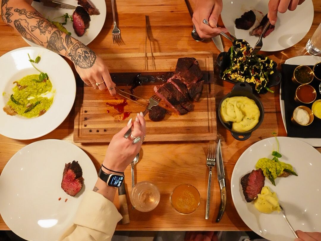 Friends sharing a premium steak dinner with sides like mashed potatoes, grilled vegetables, and sauces in a lively, upscale setting.