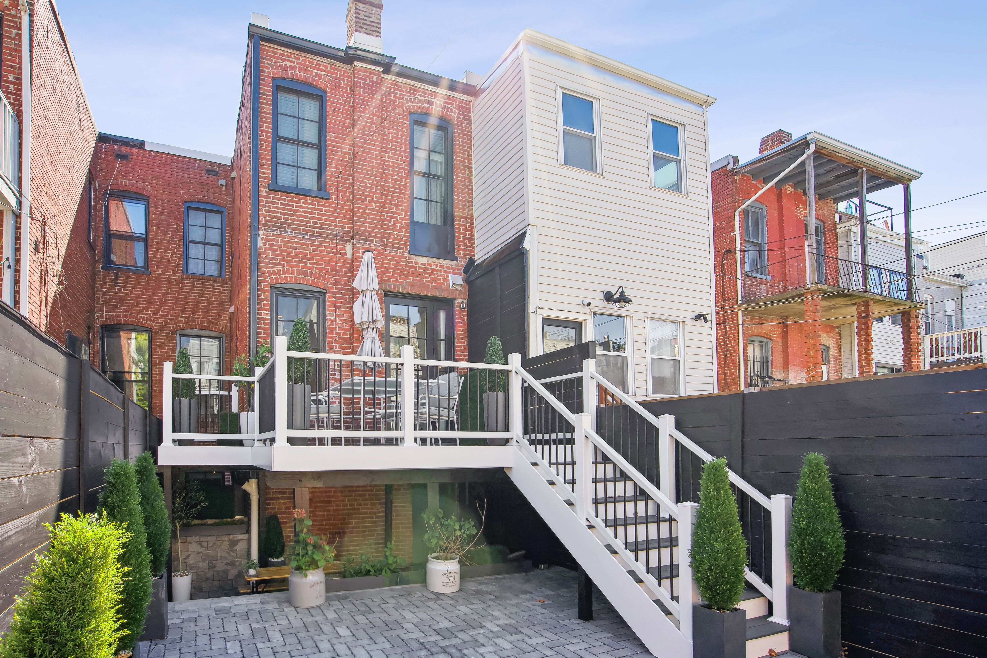 Renovated urban townhouse with a red brick and white siding exterior, featuring a raised deck, modern black-and-white staircase, and fenced-in patio with potted greenery.