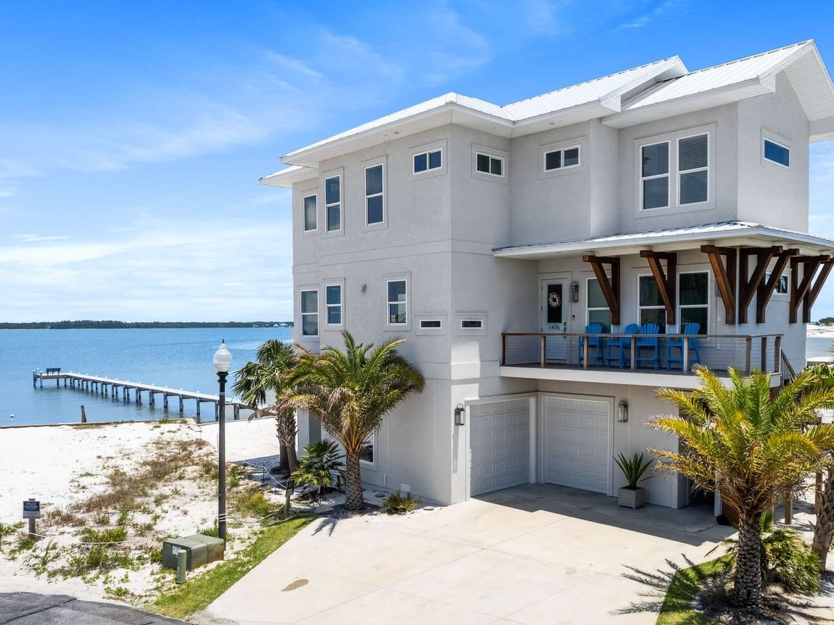 A modern white beach house with oceanfront views and a private dock stretches out into calm blue water. The home features a large balcony with bright blue chairs and palm trees lining the driveway, perfect for a peaceful coastal retreat.