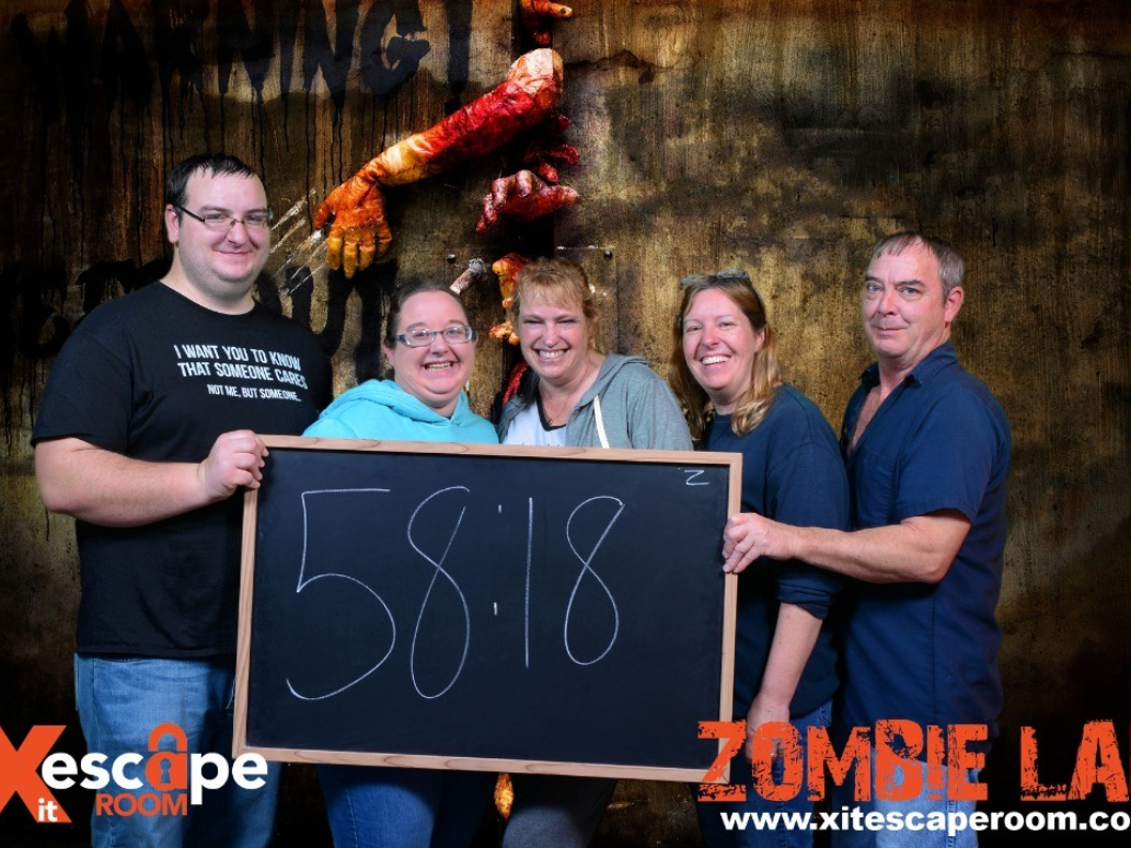 A group of five smiling people hold a chalkboard showing their escape time in front of a zombie-themed backdrop at Xscape It Room's "Zombie Lab."
