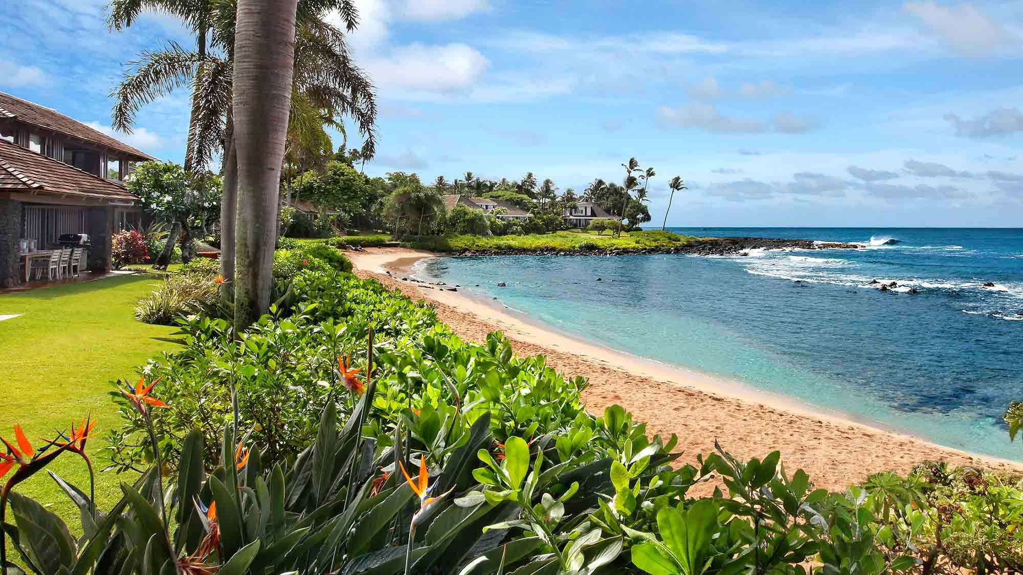 Honu Kai Villa in Kauai, Hawaii, overlooks a calm sandy cove with clear blue water and swaying palm trees. Tropical gardens and oceanfront seating create a peaceful beachfront setting. This private island villa offers a relaxing Hawaii getaway with direct beach access and stunning coastal views.