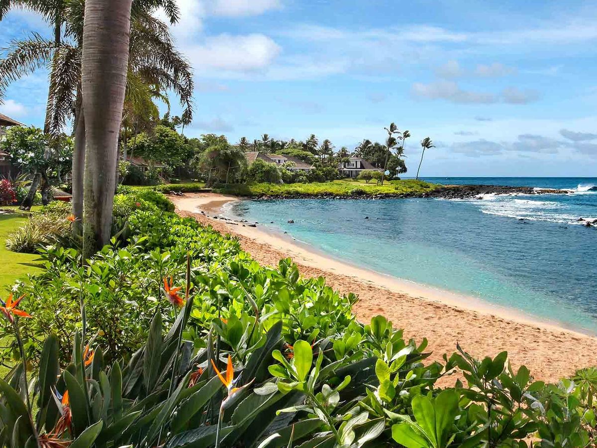 Honu Kai Villa in Kauai, Hawaii, overlooks a calm sandy cove with clear blue water and swaying palm trees. Tropical gardens and oceanfront seating create a peaceful beachfront setting. This private island villa offers a relaxing Hawaii getaway with direct beach access and stunning coastal views.