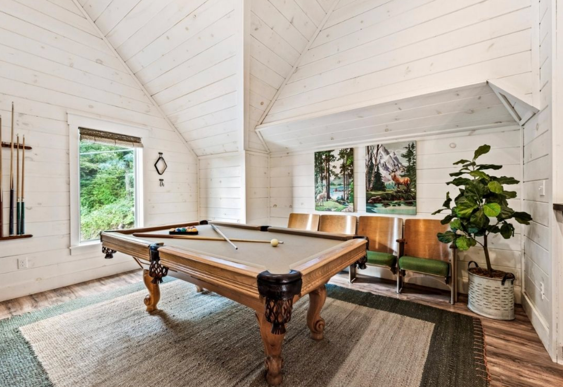 Bright, airy game room with vaulted shiplap ceilings, a pool table, cozy seating, and large windows overlooking lush greenery.