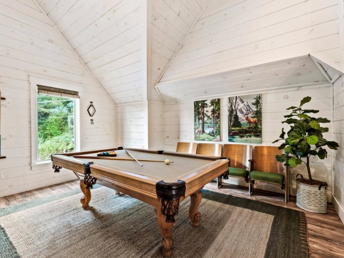 Bright, airy game room with vaulted shiplap ceilings, a pool table, cozy seating, and large windows overlooking lush greenery.