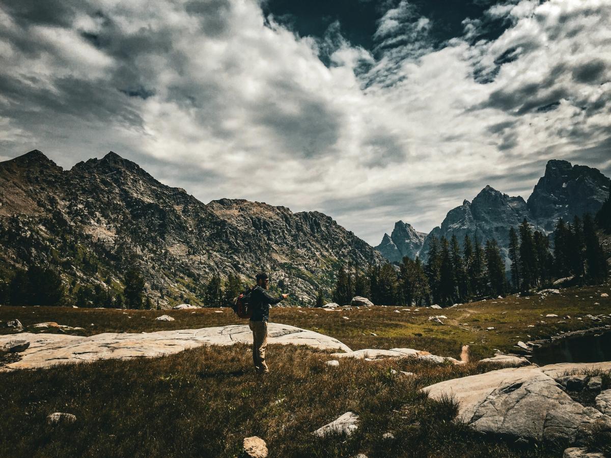 A hiker with a backpack stands in an alpine meadow surrounded by rugged mountain peaks and dramatic clouds in a remote wilderness landscape.