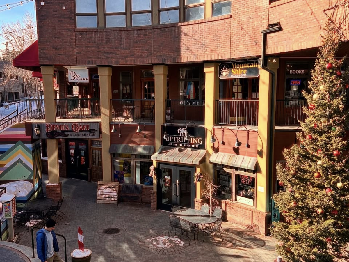 Charming brick shopping plaza with local stores, a large decorated Christmas tree, and a rainbow mural staircase, captured on a sunny winter day.