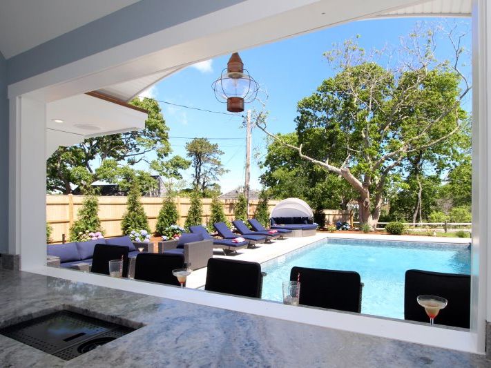 Covered outdoor bar window overlooking a private backyard pool with lounge chairs, shaded daybed, mature trees, and cocktails set along a granite counter—perfect for effortless indoor-outdoor entertaining.