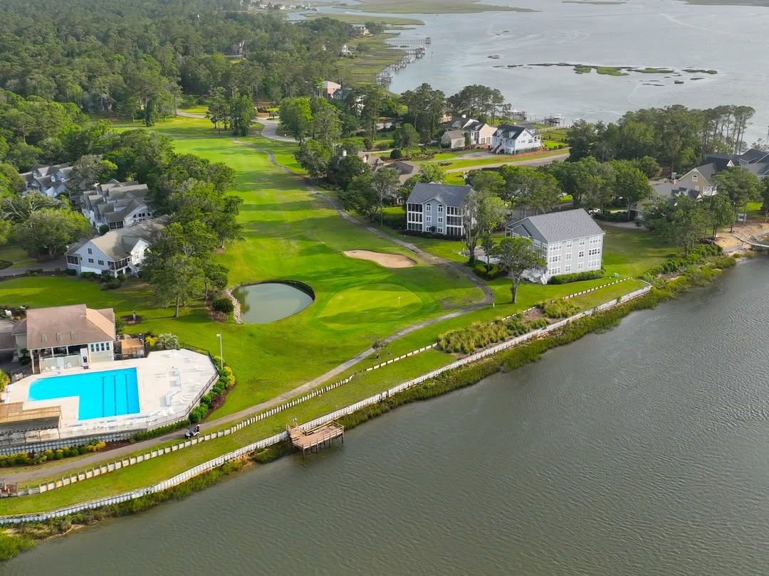 Aerial view of a peaceful golf course lined with trees and homes beside the water, with a bright pool near the clubhouse. A quiet escape in Holden Beach.