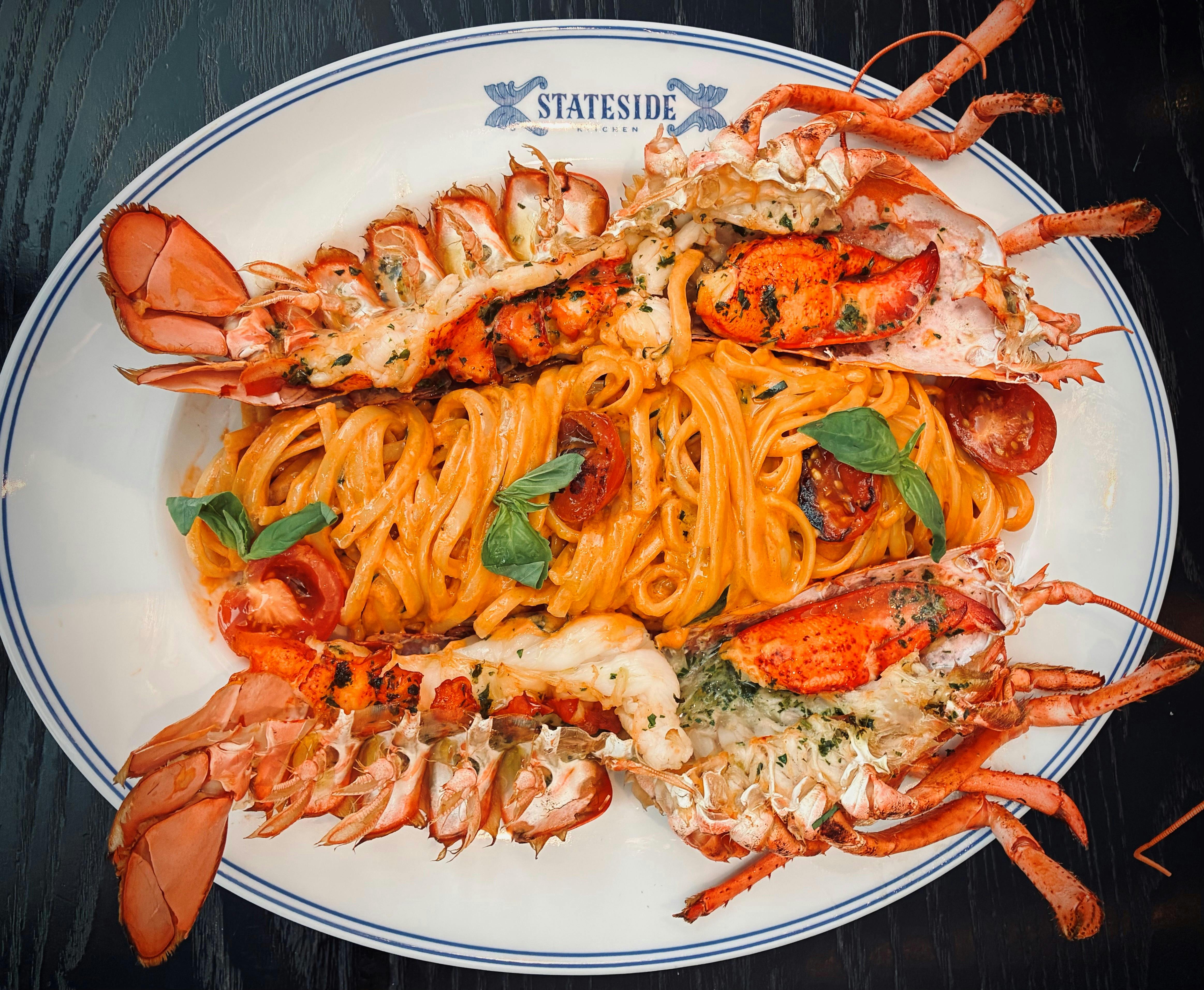 A luxurious seafood pasta dish featuring rich lobster halves atop creamy linguine with herbs and cherry tomatoes. Served on elegant white dinnerware, it’s a feast for both the eyes and the palate.