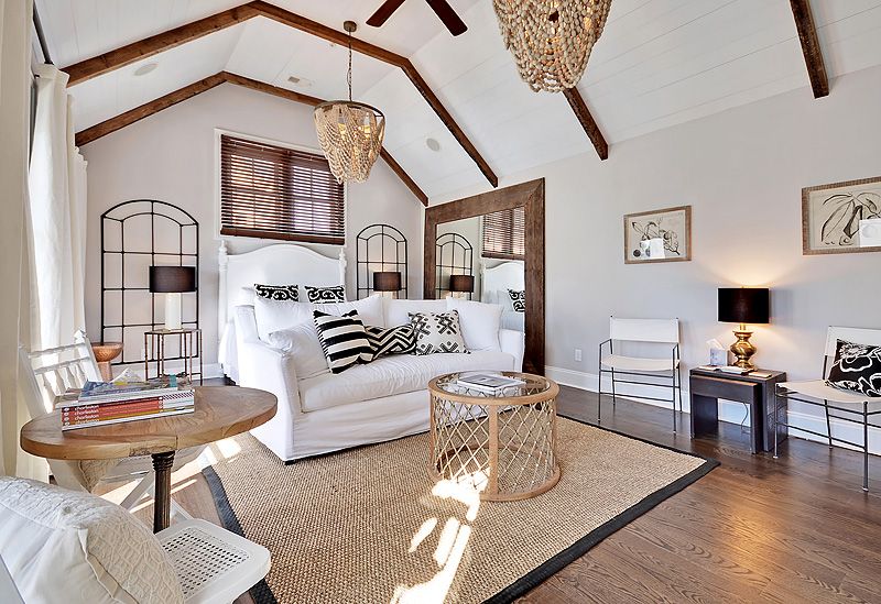 A bright, airy bedroom with vaulted ceilings and exposed wood beams features a white bed, neutral-toned furnishings, black and white patterned pillows, and natural textures like woven rugs and light fixtures.