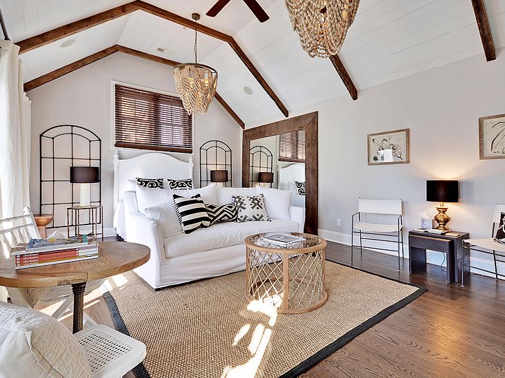 A bright, airy bedroom with vaulted ceilings and exposed wood beams features a white bed, neutral-toned furnishings, black and white patterned pillows, and natural textures like woven rugs and light fixtures.