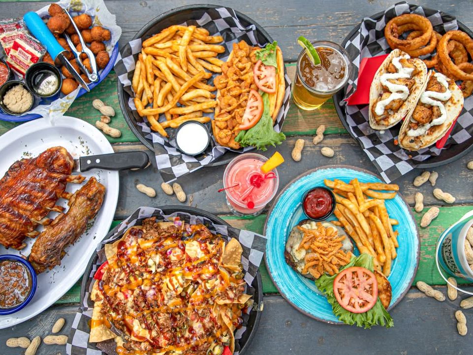 A colorful table of coastal comfort food at Boardwalk Billy’s in North Myrtle Beach, with BBQ ribs, nachos, fried shrimp, fish tacos, burgers, and frosty cocktails.