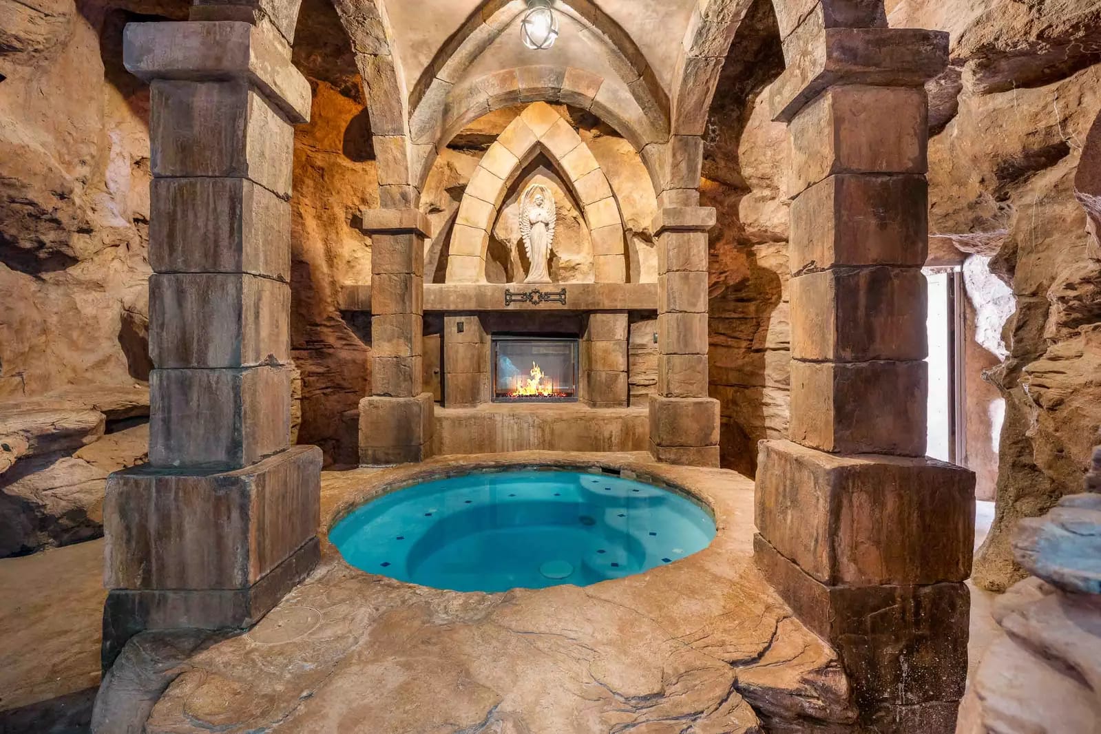 Stone grotto-style spa with a round hot tub, gothic arches, and a fireplace set into a carved rock wall.