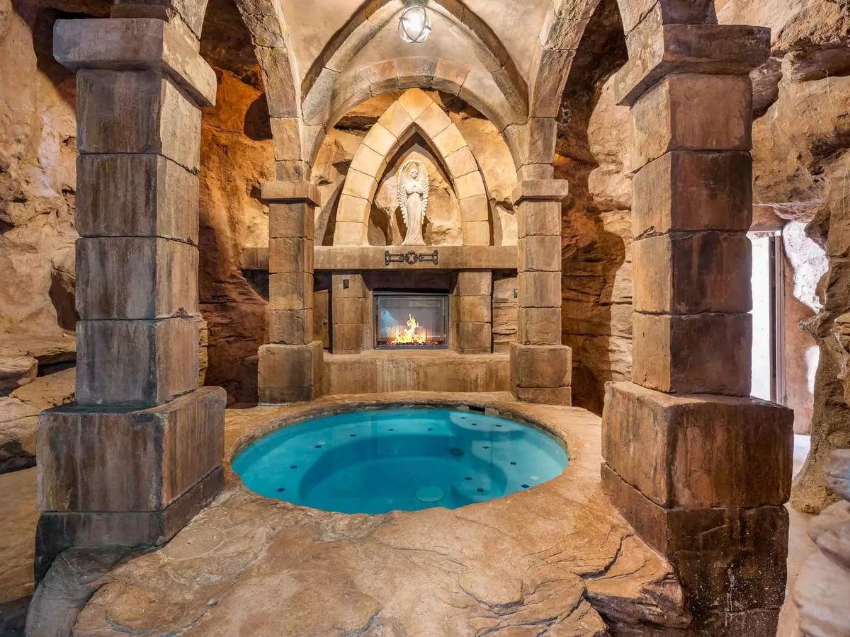 Stone grotto-style spa with a round hot tub, gothic arches, and a fireplace set into a carved rock wall.