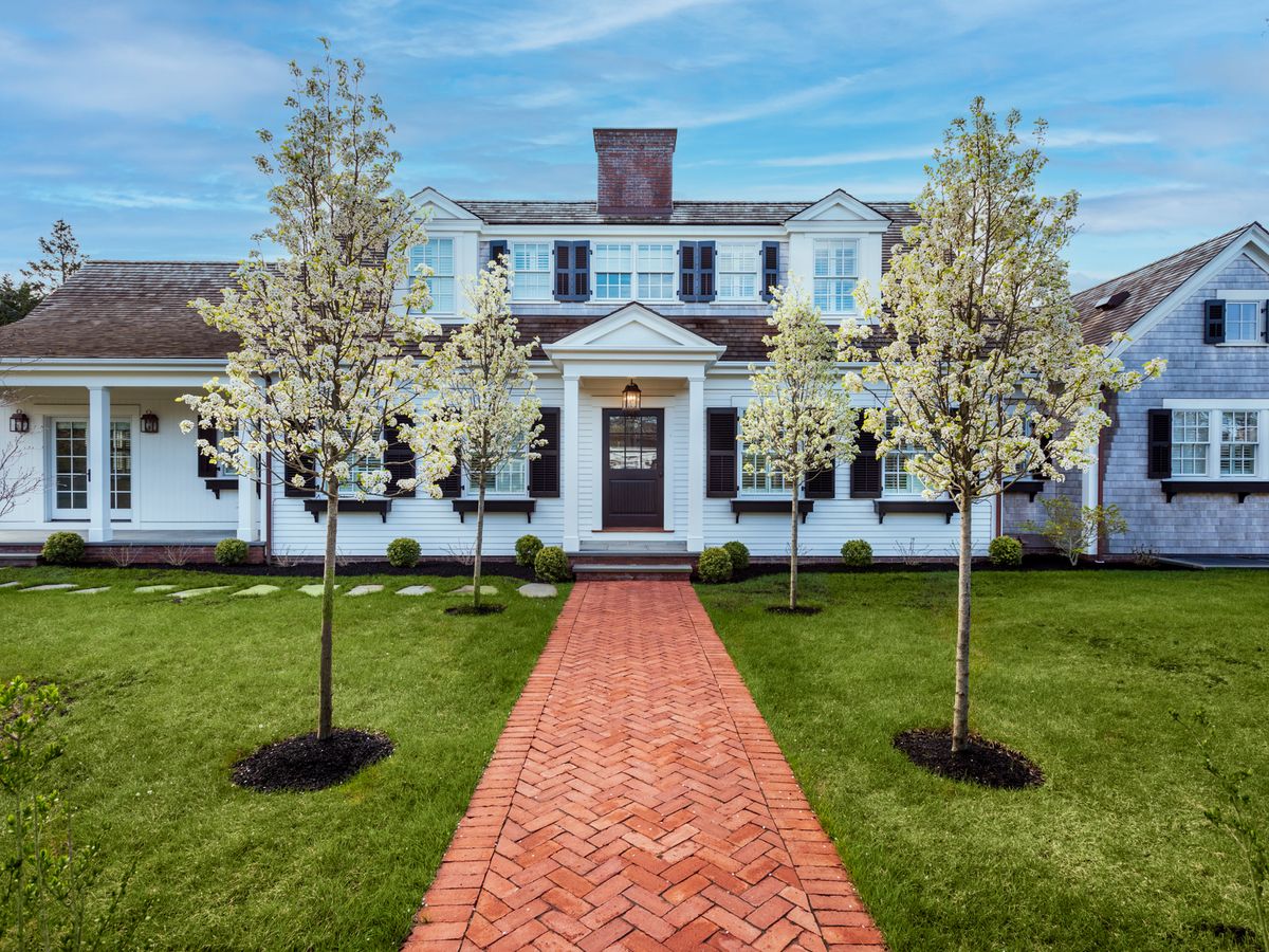 A stately coastal home with white siding, black shutters, and a red brick walkway is framed by blooming trees and a perfectly manicured lawn under a bright blue sky.
