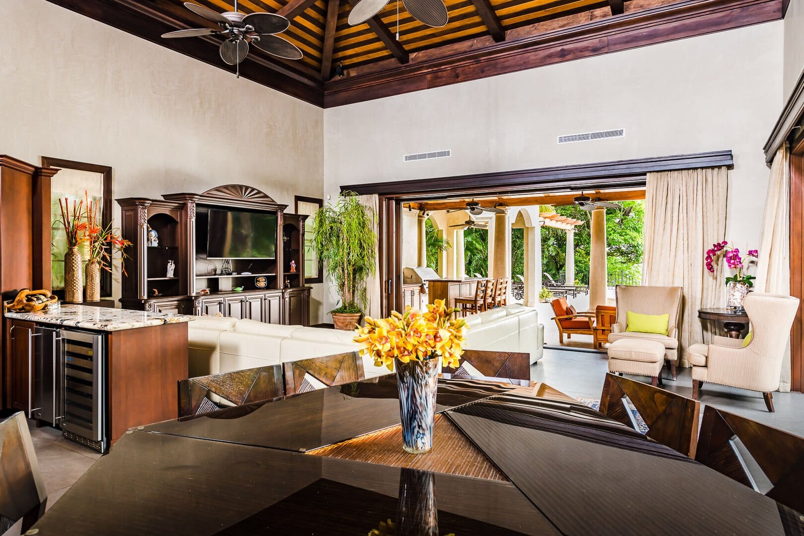 Spacious living room with vaulted wood-beam ceilings, elegant dark wood furniture, and sliding doors that open to a covered outdoor patio.