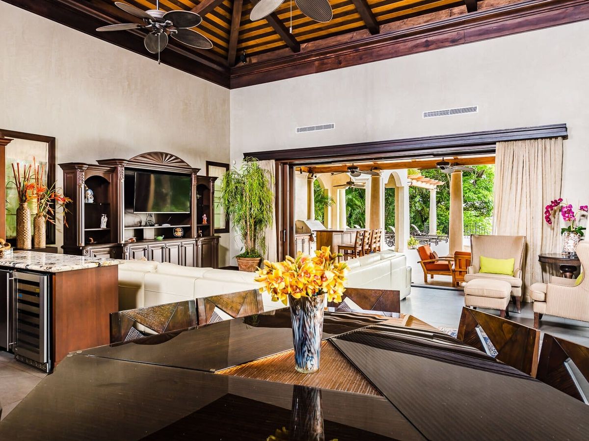 Spacious living room with vaulted wood-beam ceilings, elegant dark wood furniture, and sliding doors that open to a covered outdoor patio.