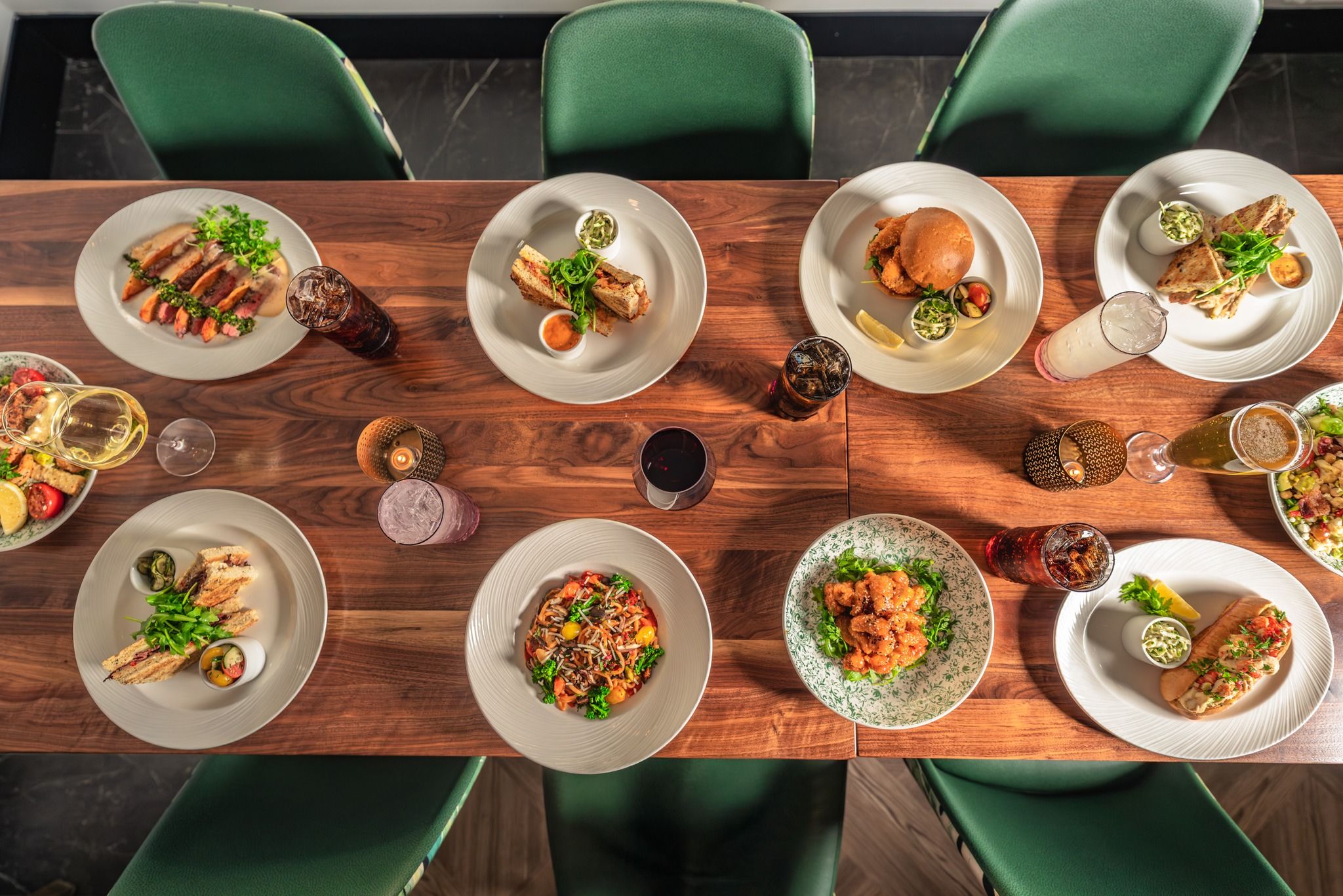 A long wooden table is filled with colorful plates of pasta, sandwiches, salads, and crispy appetizers, creating a warm and inviting dining scene. Each dish looks fresh and vibrant, making it easy to imagine sharing a meal with friends. The mix of drinks and candlelight adds a cozy, modern touch to the setting.