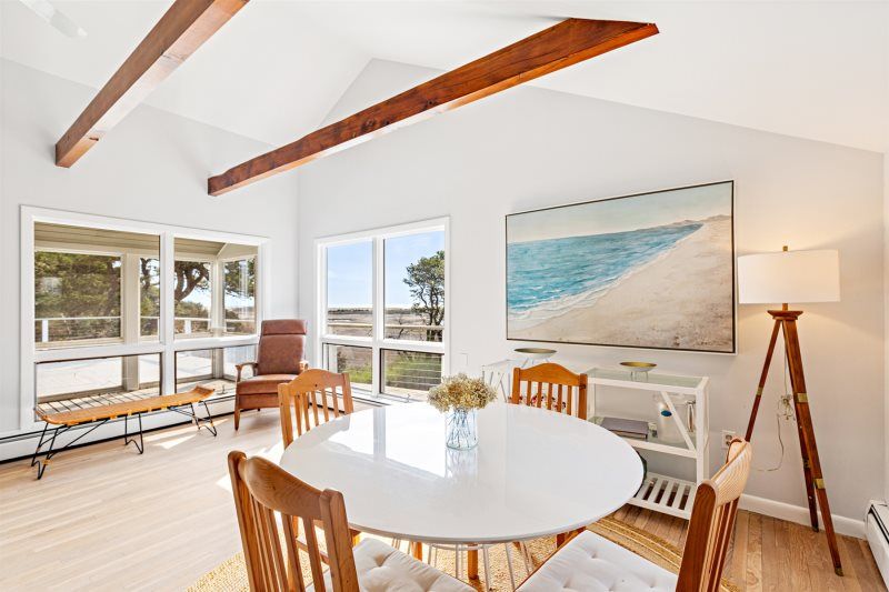 Bright coastal dining area with vaulted ceilings and exposed wood beams, featuring a round white table, natural wood chairs, beach artwork, and large windows overlooking a serene waterfront view.