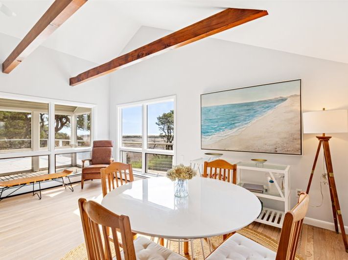 Bright coastal dining area with vaulted ceilings and exposed wood beams, featuring a round white table, natural wood chairs, beach artwork, and large windows overlooking a serene waterfront view.
