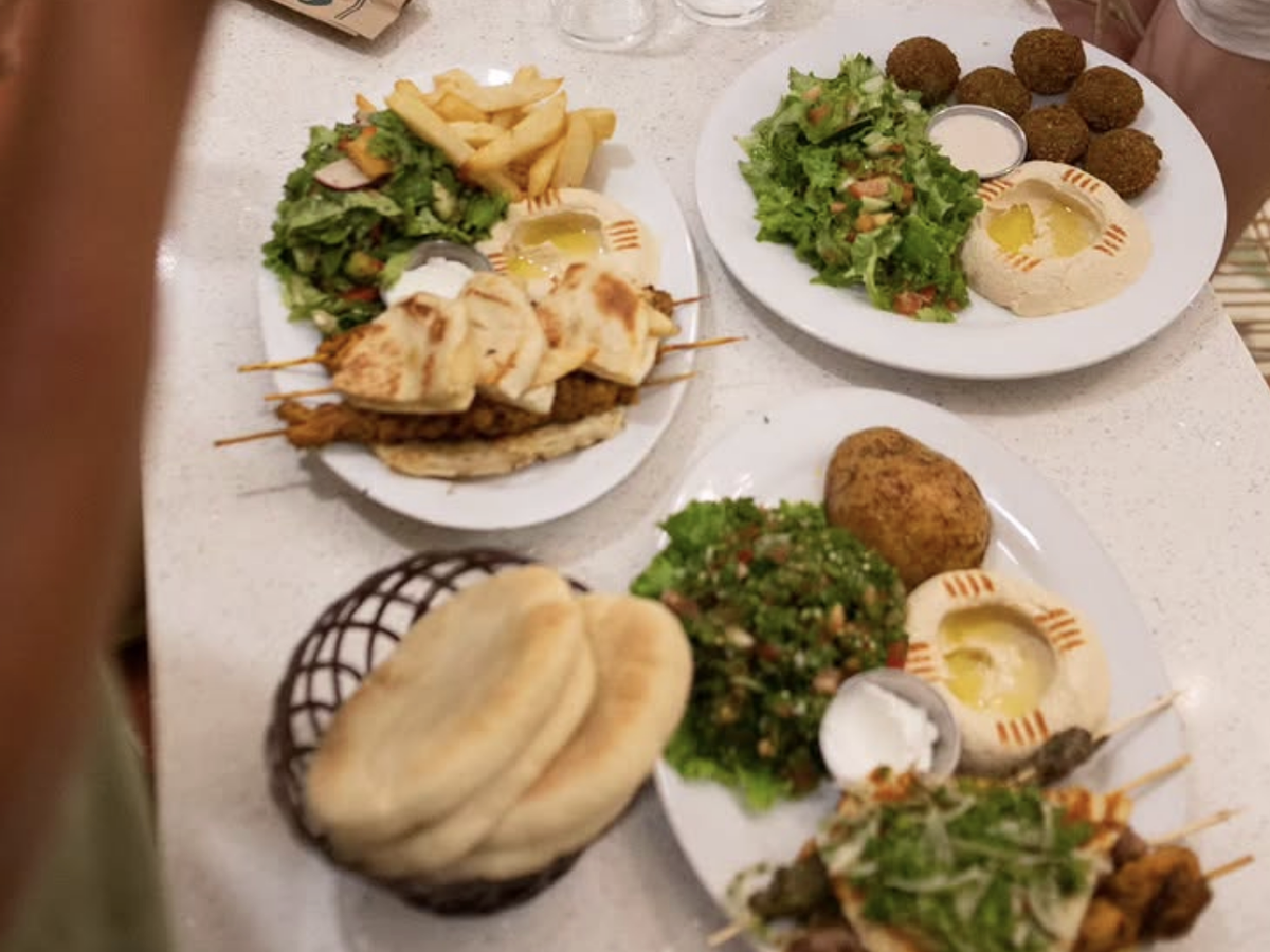 Plates of Middle Eastern food featuring skewers, hummus, falafel, fresh salad, fries, pita bread, and tabbouleh on a white table