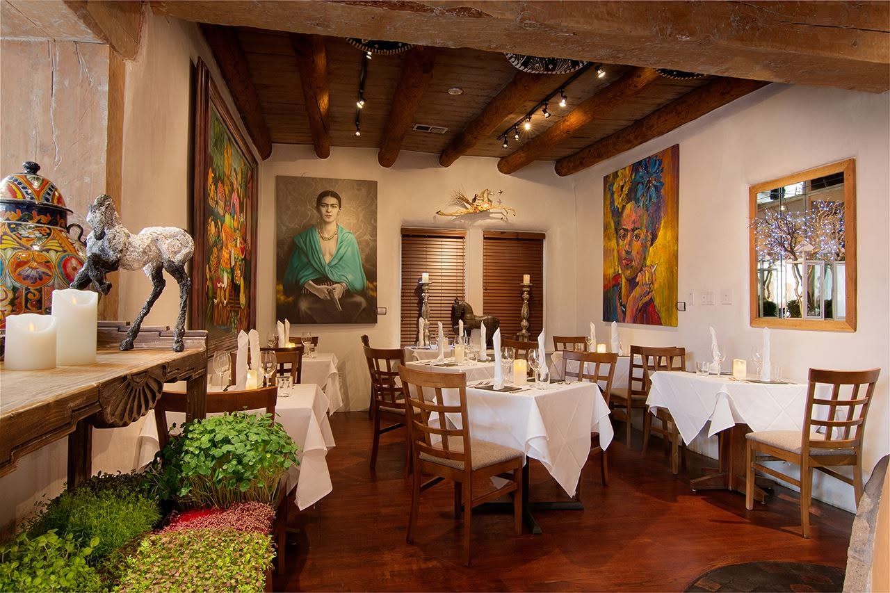 A warmly lit dining room features exposed viga ceiling beams, white adobe walls, and crisp white-linen tables set with candles and folded napkins. Vivid portraits of Frida Kahlo and sculptural artwork give the intimate space a distinctly artistic Southwestern character. 